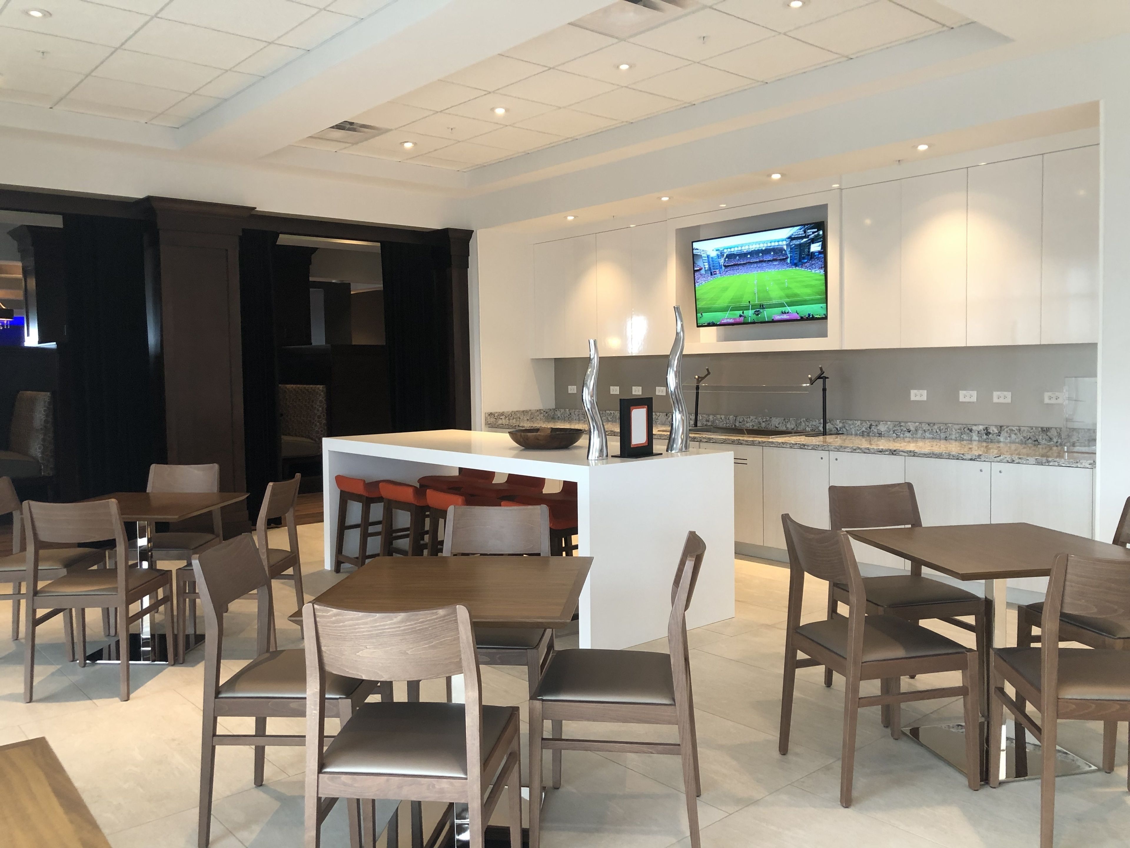 A modern hotel dining area with a central island, bar stools, and a television mounted above the kitchen counter.