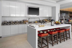 A modern kitchen with white cabinets, a large island with bar stools, and a flat-screen TV mounted above the island. - Hilton Garden Inn Denver/Thornton (Thornton)