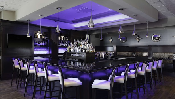 A bar area with a long counter, multiple bar stools, and a well-stocked shelf with bottles. - Hilton Garden Inn Denver/Thornton (Thornton)
