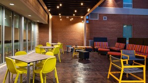 Un área de estar interior con sillas y mesas amarillas, pared de ladrillo y techo con luces.