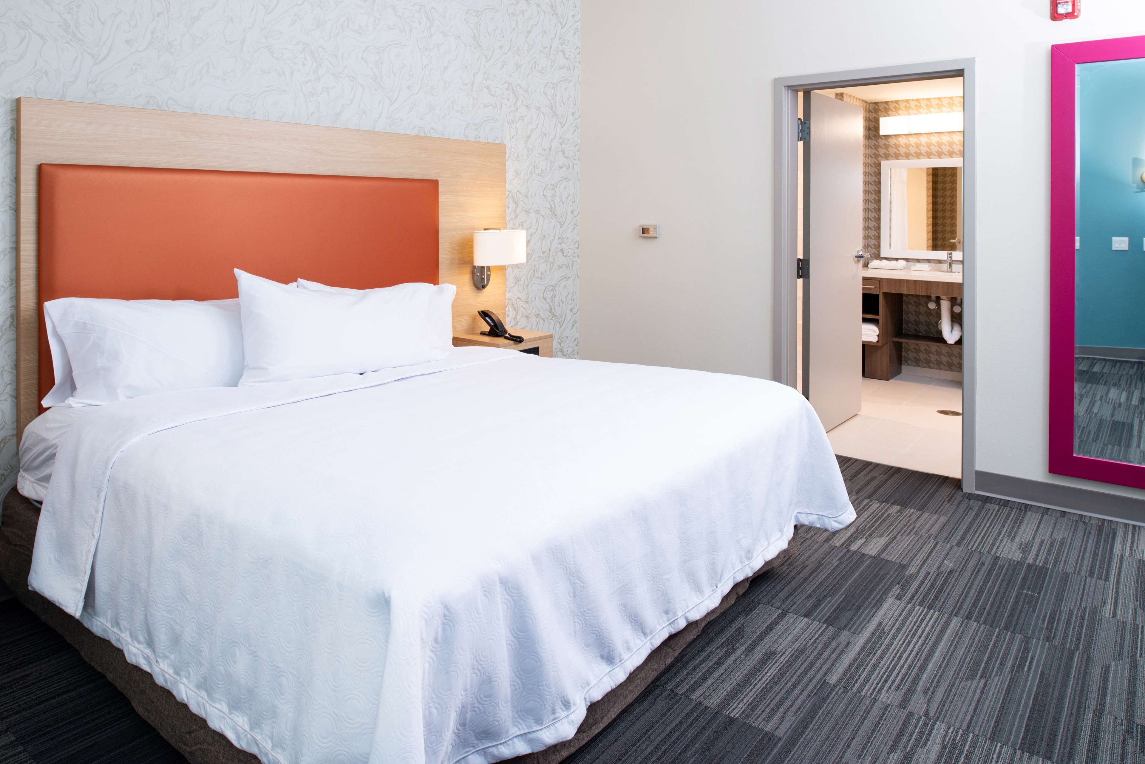 Une chambre d’hôtel avec un grand lit, une table de chevet avec une lampe et une salle de bain visible à travers une porte ouverte.