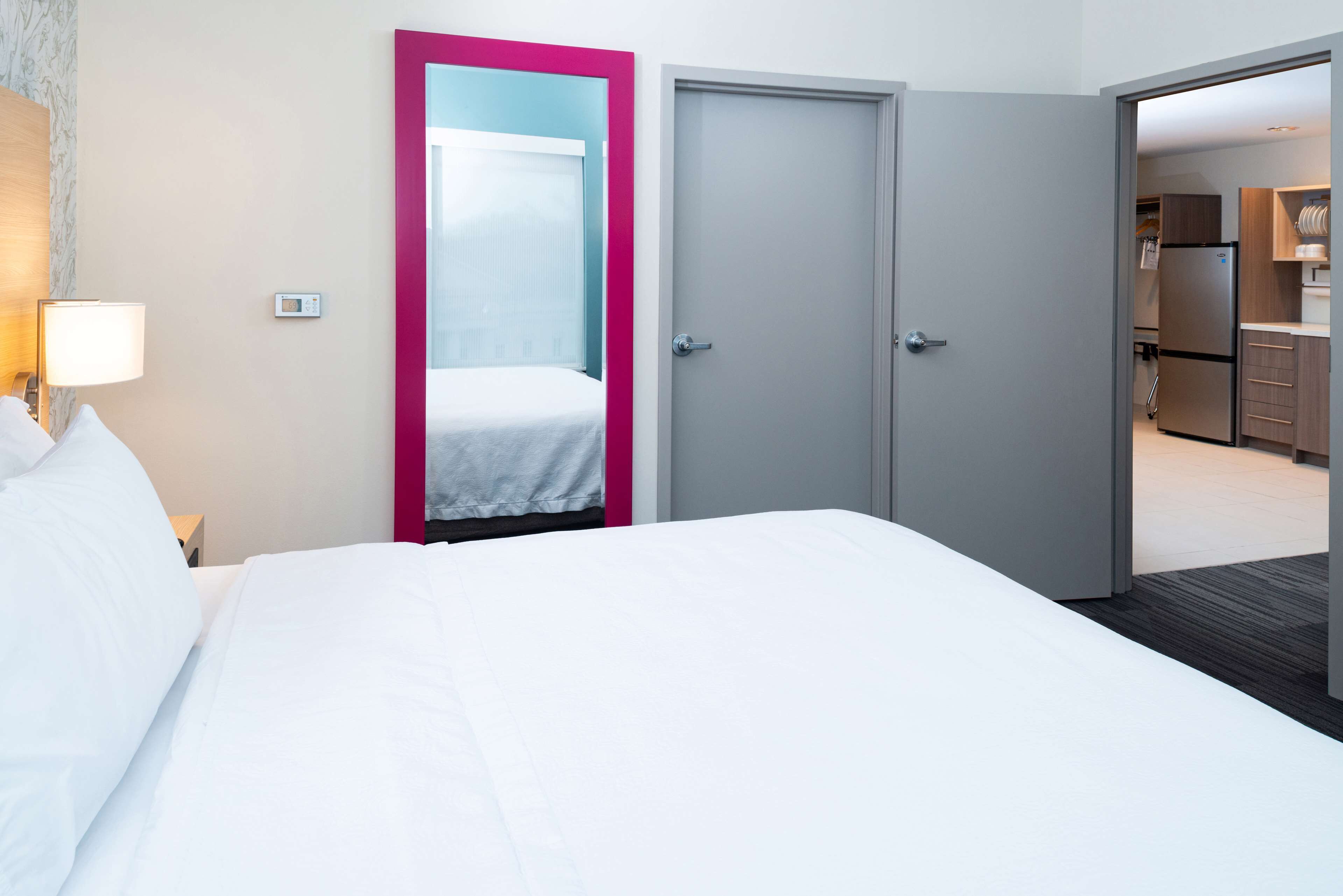 Une chambre d’hôtel comprenant un lit, une table de chevet, une lampe, un miroir et une porte donnant sur une petite cuisine.