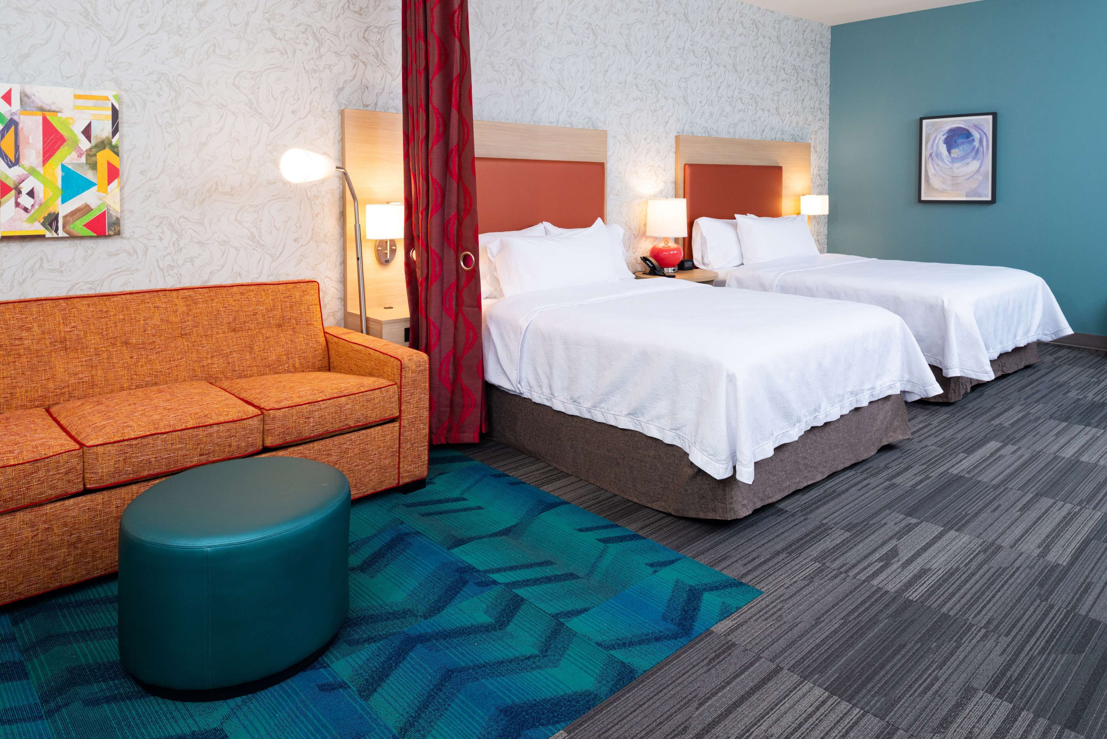 Une chambre d’hôtel avec deux lits, un canapé, un pouf rond et un tapis à motifs.