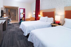 Quarto de hotel com duas camas, uma escrivaninha, uma cadeira e uma pequena mesa com abajur.