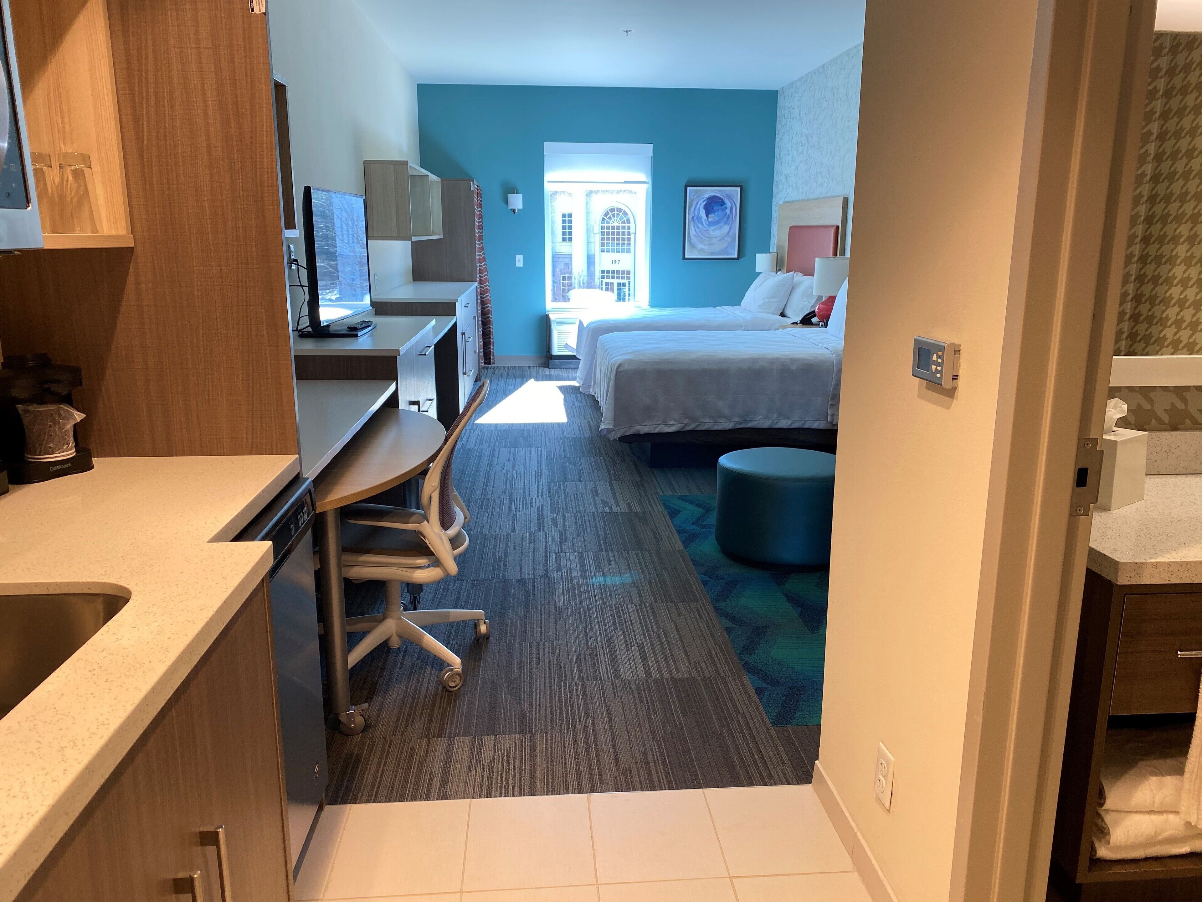 A hotel room with a kitchenette, a bed, a desk, and a bathroom.