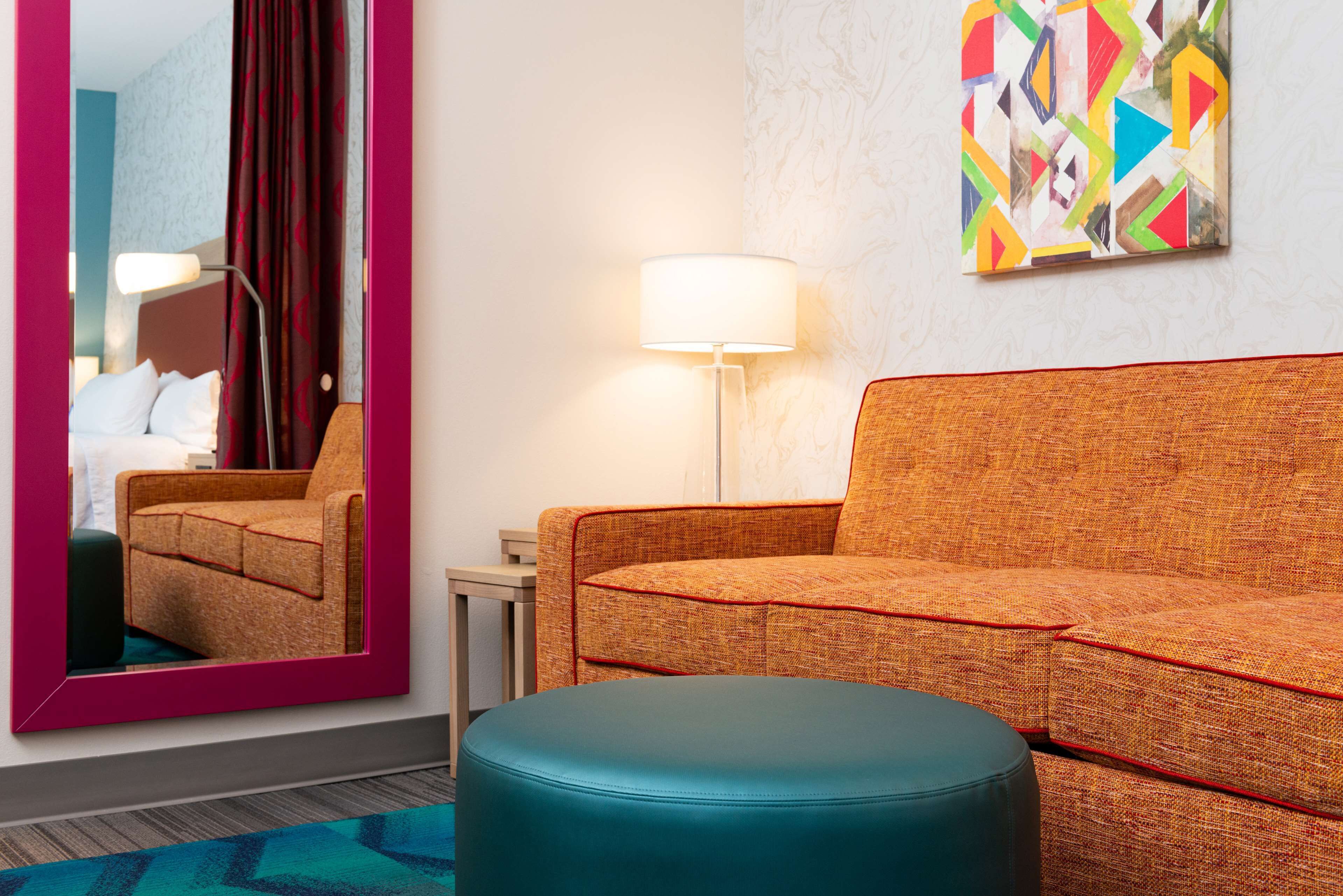 A hotel room with a teal ottoman, a patterned orange sofa, a mirror with a pink frame, and a colorful abstract painting on the wall.