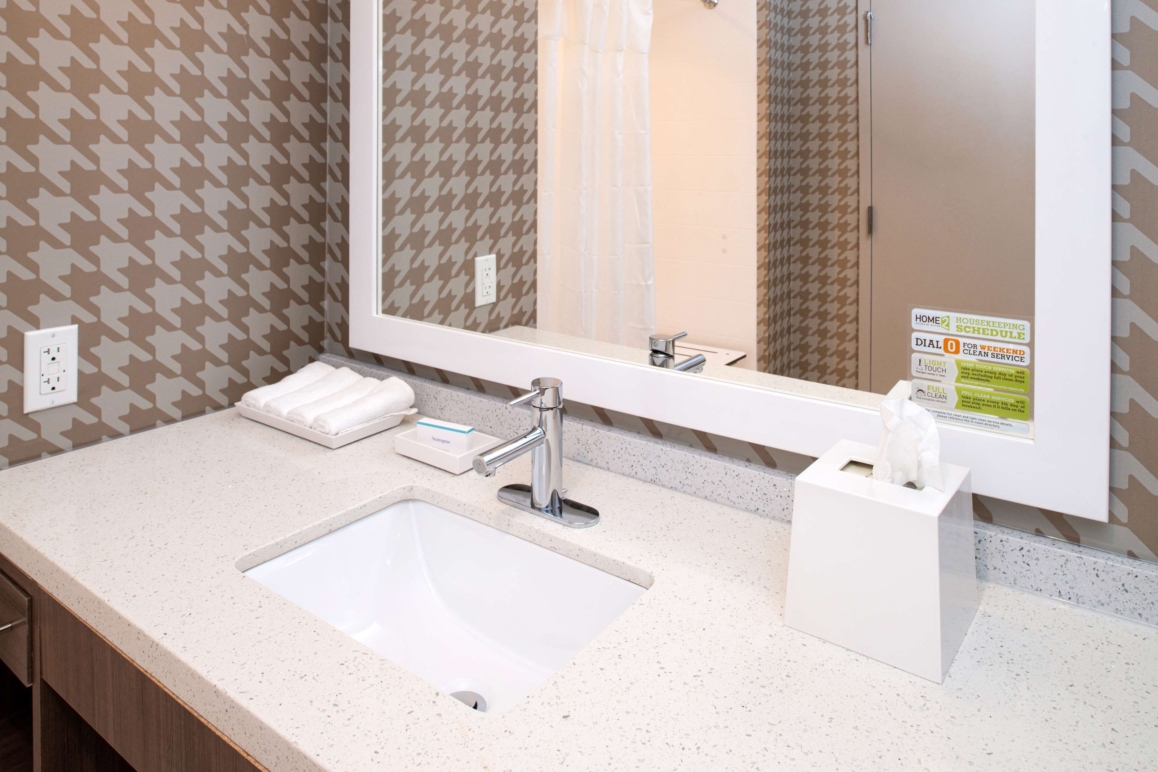 A bathroom with a white sink, a mirror, and a towel rack. There is a sign on the wall indicating housekeeping schedules.
