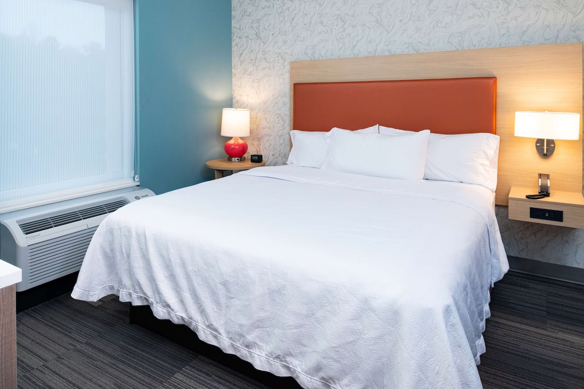 A hotel room with a large bed, two bedside tables with lamps, a heater, and a window with blinds.