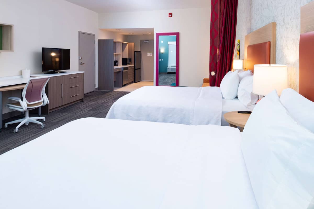 A hotel room with two beds, a desk with a chair, a television, and a small kitchen area.