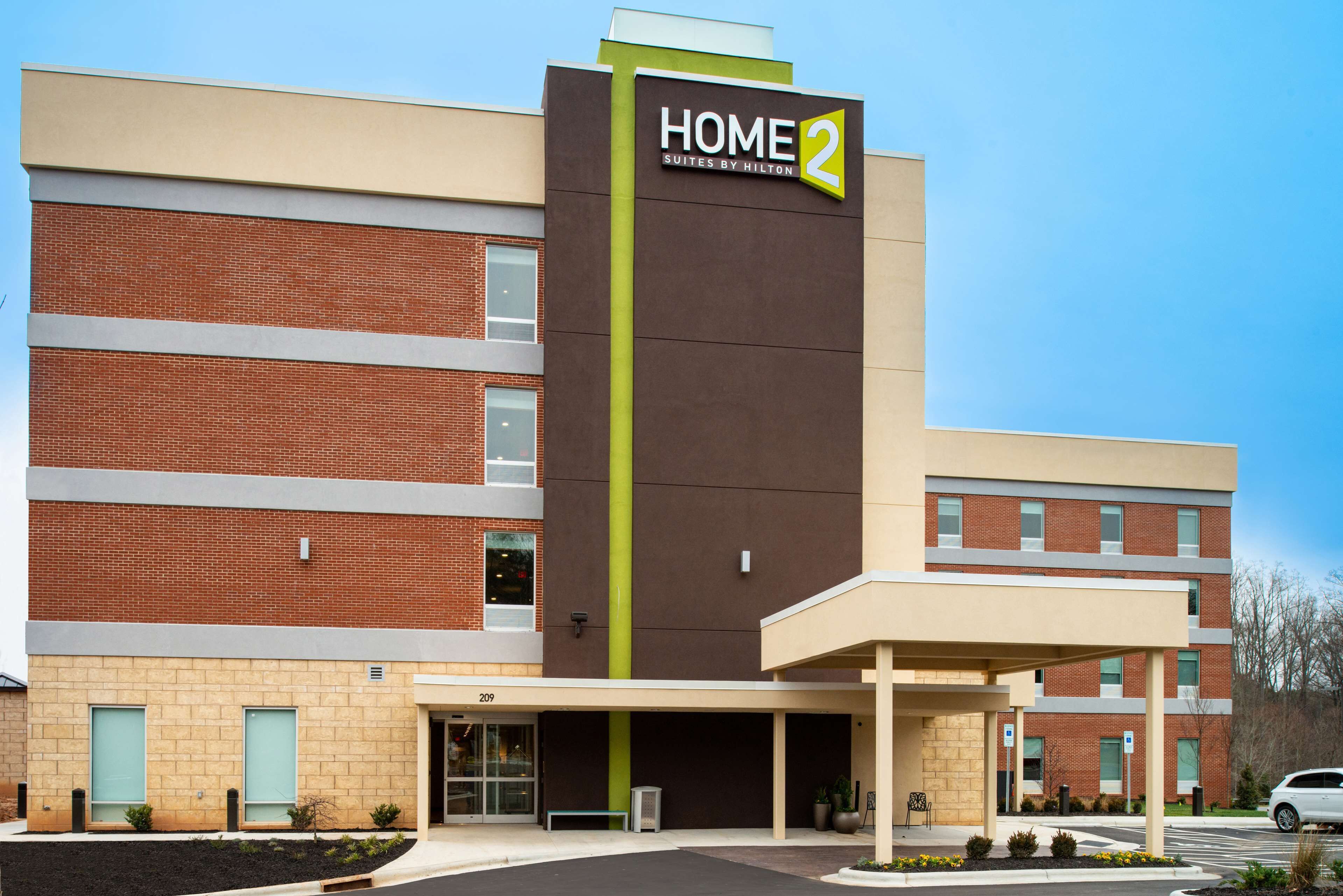 Ein modernes Hotel mit einem auffälligen Schild, das "HOME 2 Suites by Hilton" zeigt, einer Ziegel-Fassade und einem überdachten Eingangsbereich.