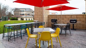 Un patio exterior con mesas y sillas, sombrillas rojas y una parrilla.