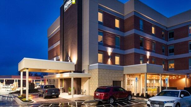 Un hotel de varios pisos con un llamativo letrero “HOME2”, una zona de estacionamiento techada y una entrada visible con asientos al aire libre.