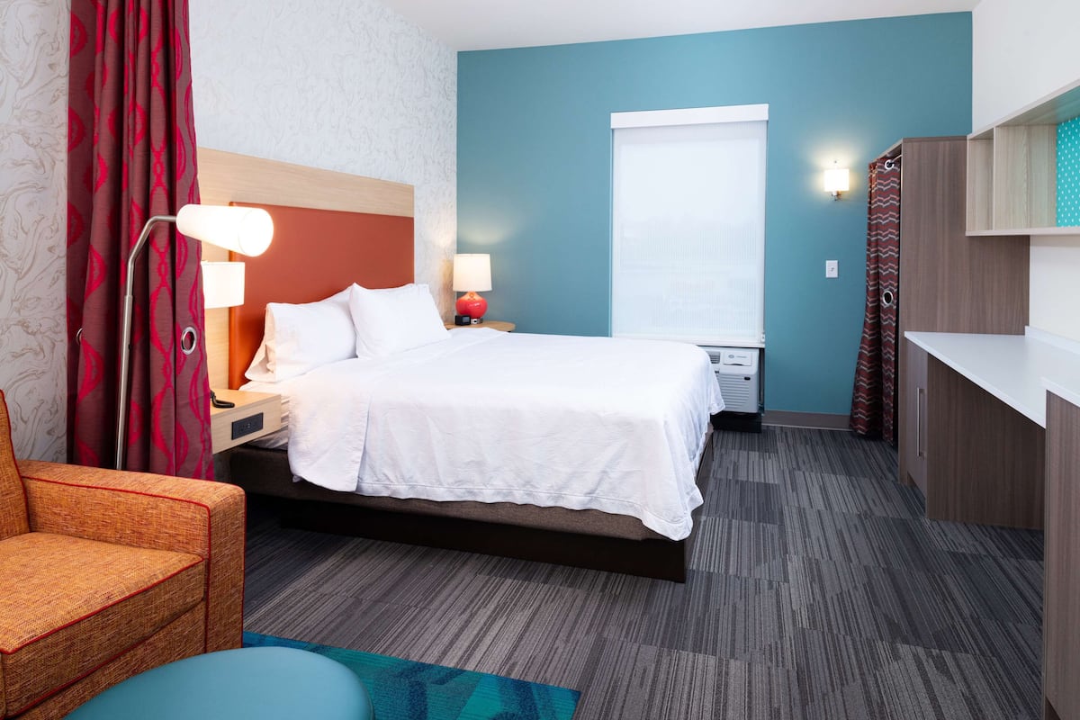 A hotel room with a bed, a desk, a chair, a lamp, a window, and a shower area.