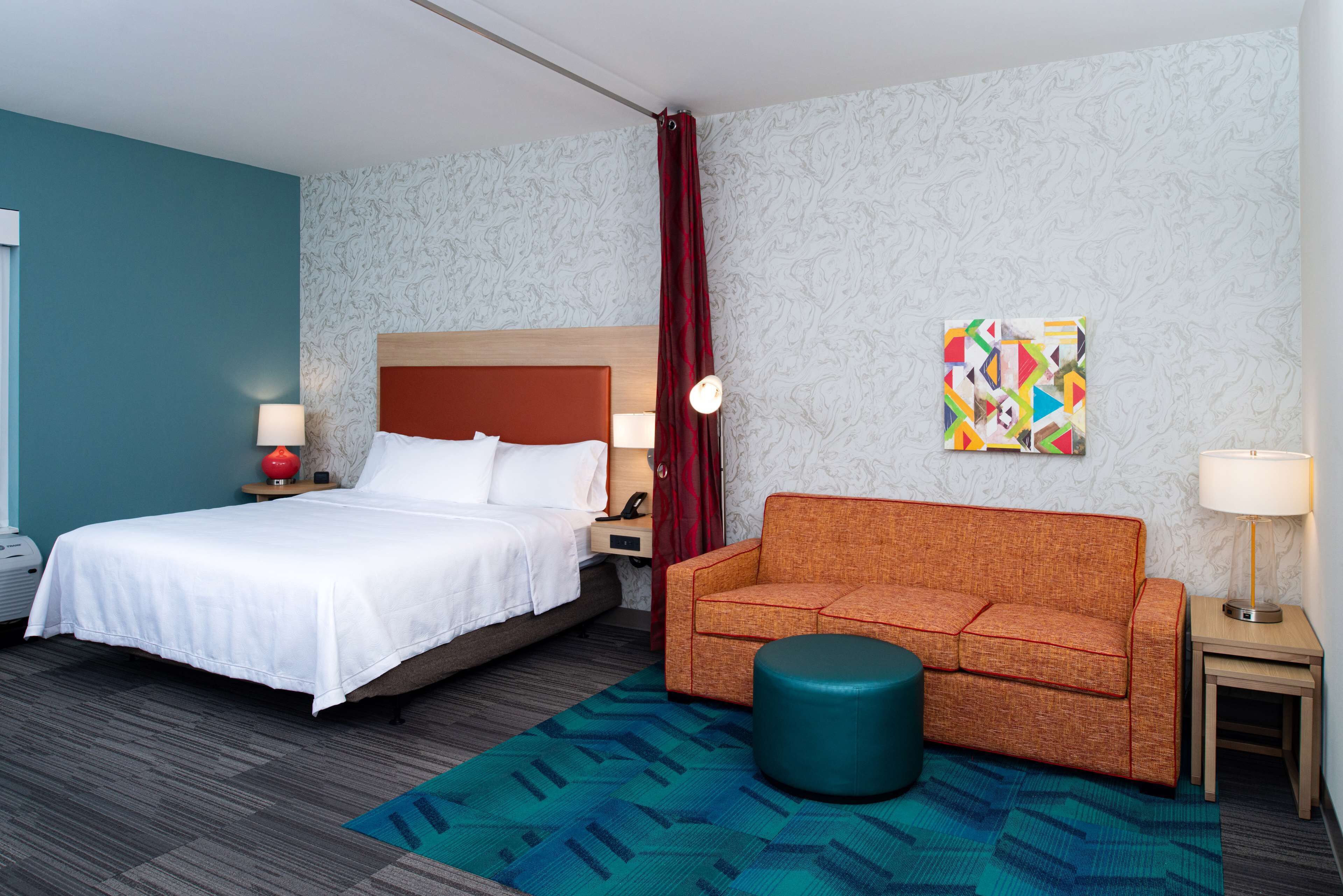 A hotel room with a bed, a sofa, a small ottoman, a nightstand, a lamp, and a wall art piece.