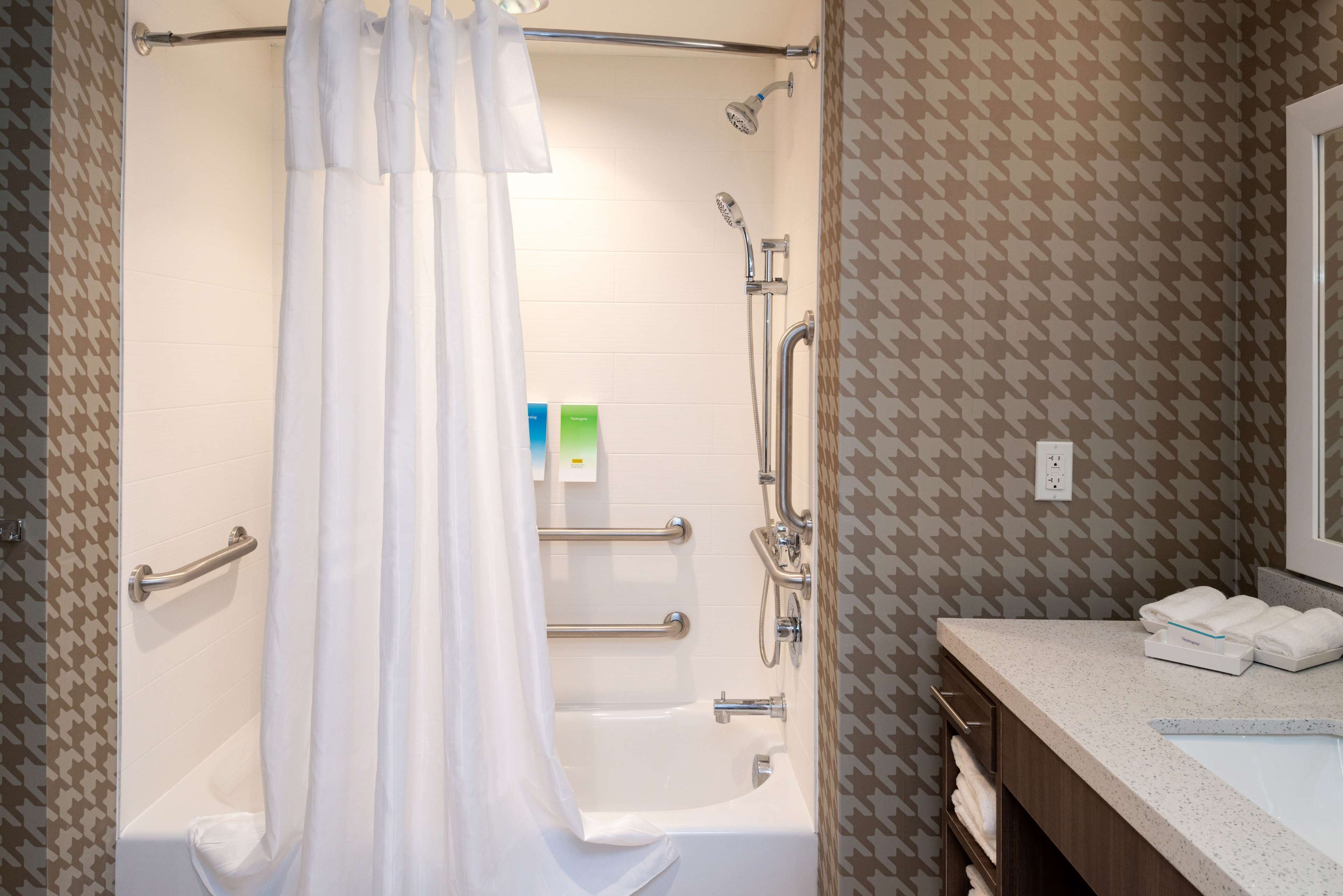 A bathroom with a bathtub, shower, and handrails. There are towels on the counter and a mirror.