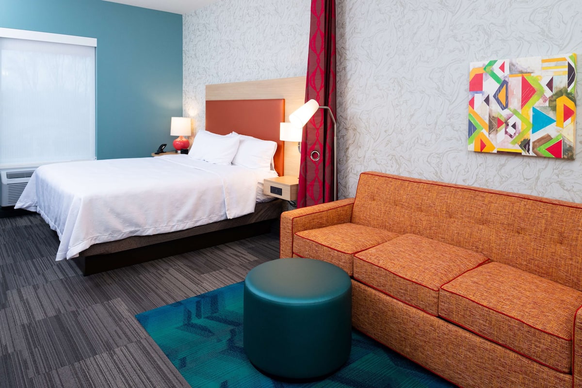 A hotel room with a bed, a sofa, a round ottoman, a lamp, and a colorful abstract painting on the wall.