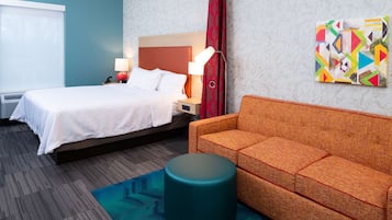 A hotel room with a bed, a sofa, a round ottoman, a lamp, and a colorful abstract painting on the wall.
