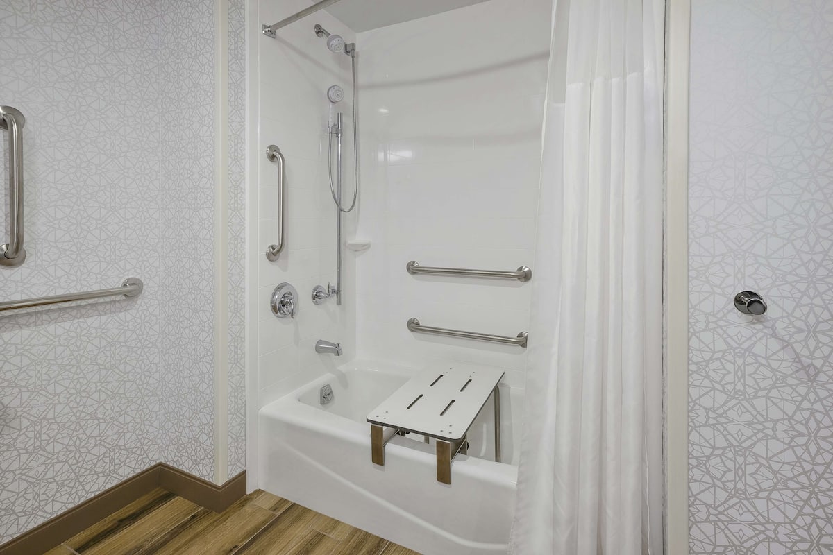 Un baño moderno con cabina de ducha, barras de apoyo y un tapete de baño de madera.