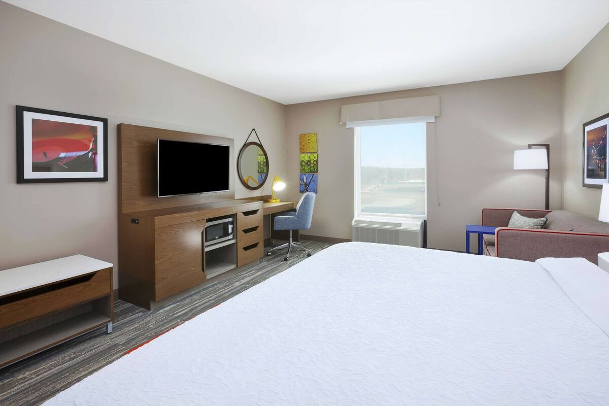 Habitación de hotel con cama, escritorio con computadora, televisión, silla, lámpara y ventana con vistas.