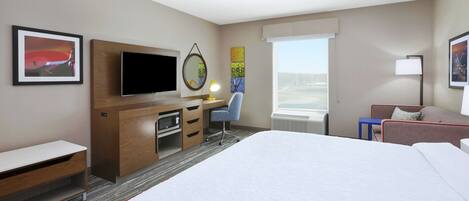 Habitación de hotel con cama, escritorio con computadora, televisión, silla, lámpara y ventana con vistas.