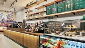 Interieur van een koffiezaak, met een houten toonbank, een koelvitrine met drankjes en schappen vol etenswaren.