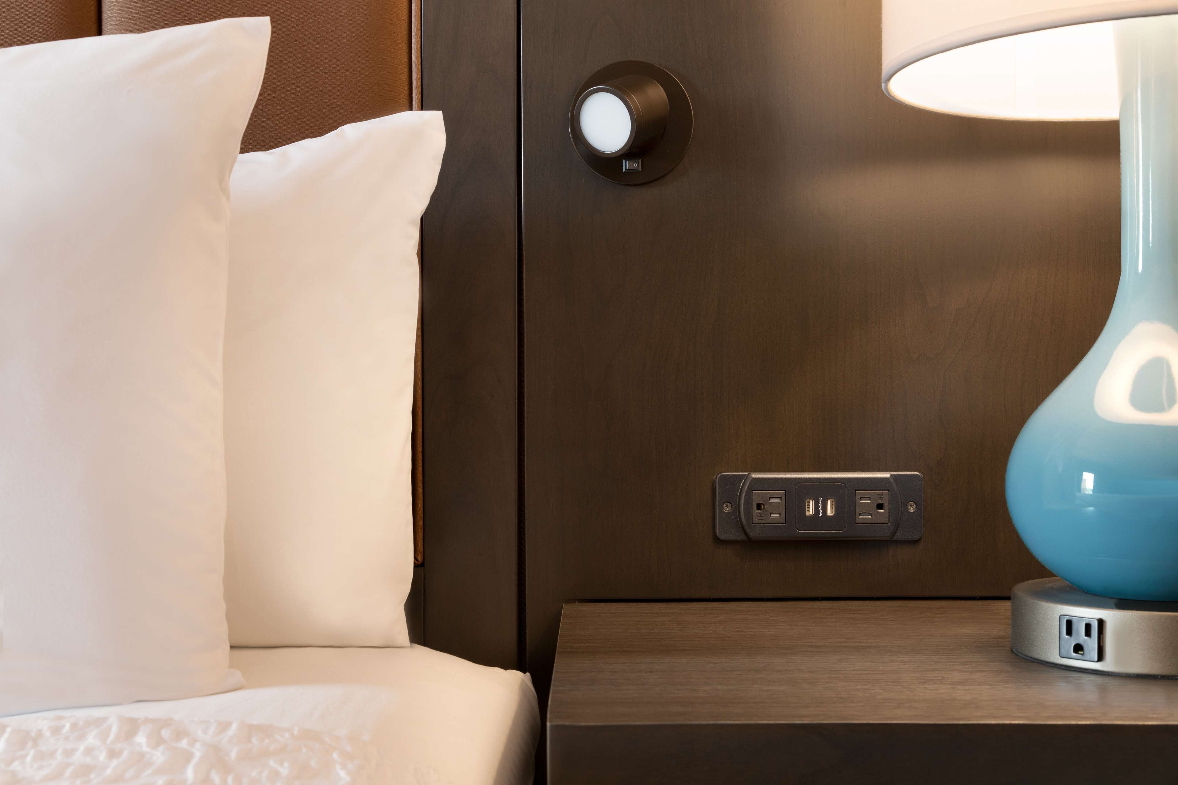 A neatly made bed with white pillows, a bedside lamp, and a control panel with two USB ports.