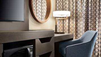 A modern hotel room with a flat-screen TV, a round mirror, a desk with a lamp, and a blue chair.