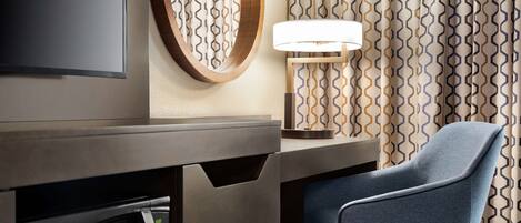 A modern hotel room with a flat-screen TV, a round mirror, a desk with a lamp, and a blue chair.