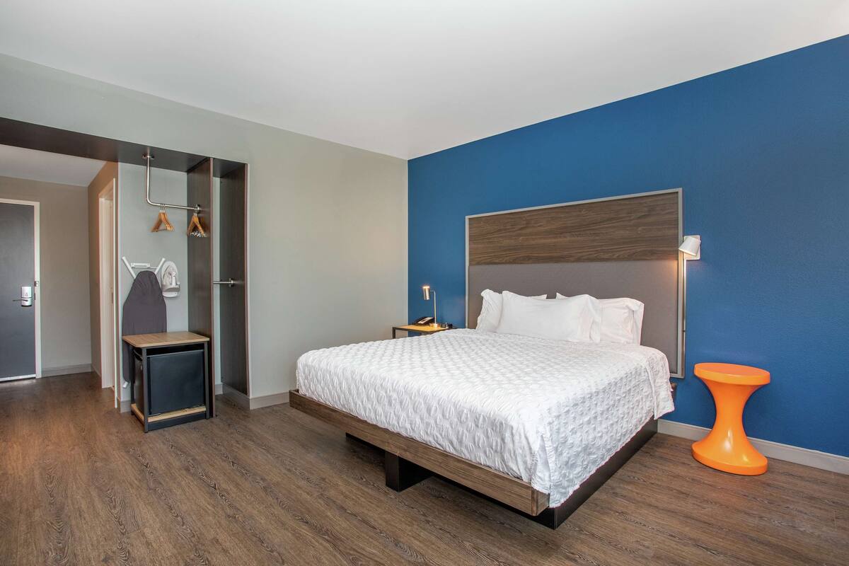 A bedroom with a bed, a bedside table, a nightstand, a hanging coat rack, and a small orange stool.