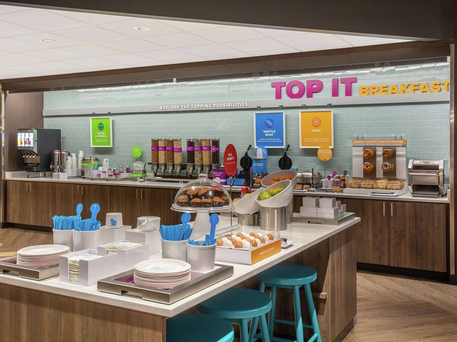 Un área de desayuno con un letrero que dice "TOP IT BREAKFAST" y diversas opciones de comida como waffles, huevos y muffins.