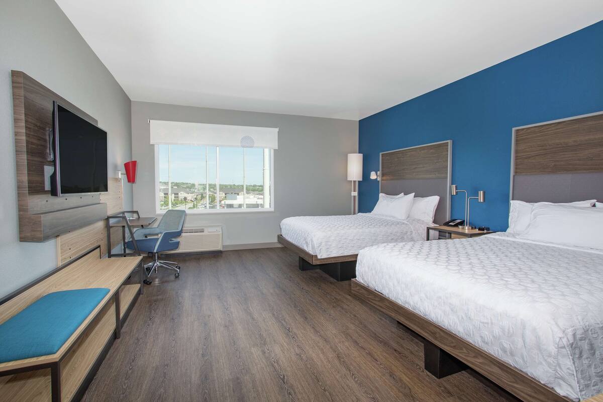 A hotel room with two beds, a desk with a chair, a TV, and a large window offering a city view.