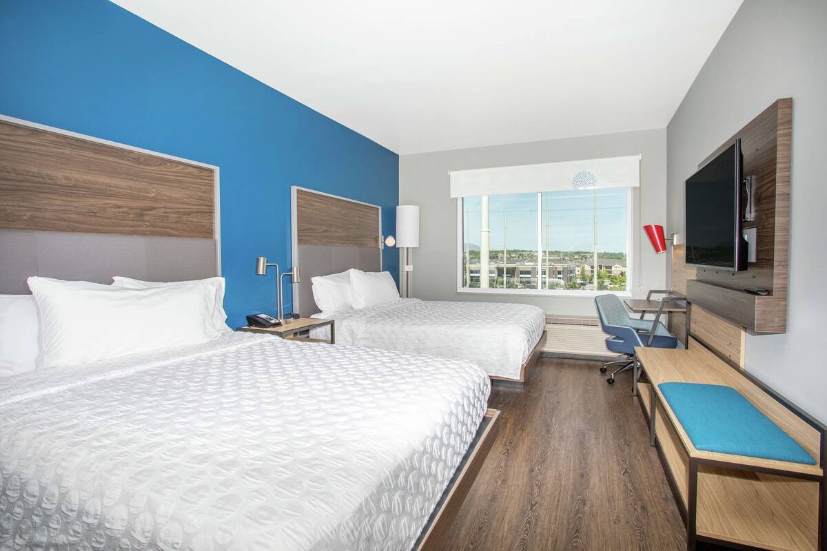 A hotel room with two beds, a desk, a TV, and a view of the city.