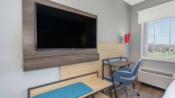 Une chambre d’hôtel dotée d’un grand téléviseur à écran plat fixé au mur, d’un bureau avec une chaise, d’un banc et d’une fenêtre donnant sur des immeubles.