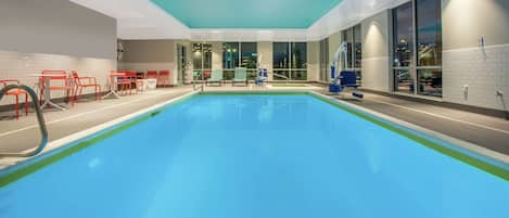 An indoor swimming pool with clear blue water, surrounded by red chairs and a blue pool lift.
