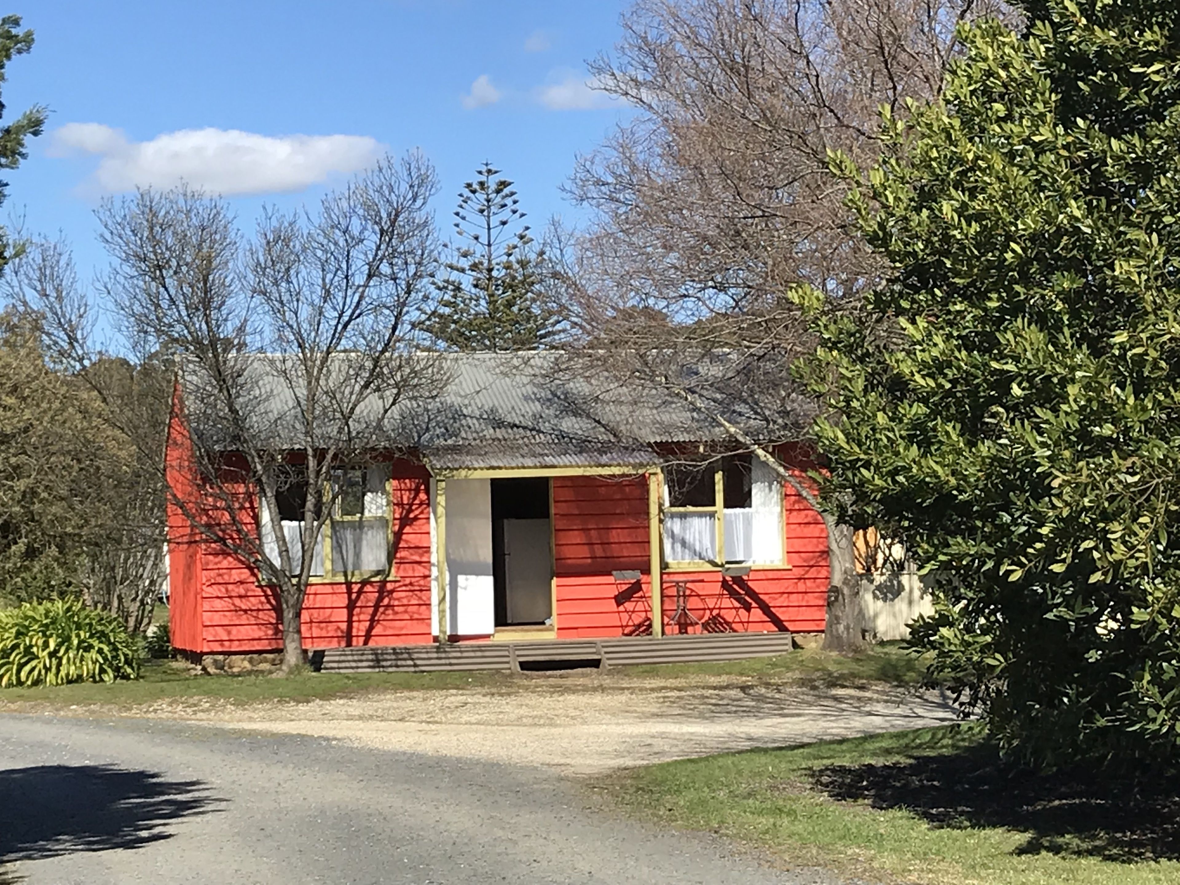 Une petite maison rouge à un étage, dotée d’un porche à l’avant, entourée d’arbres et sous un ciel dégagé.
