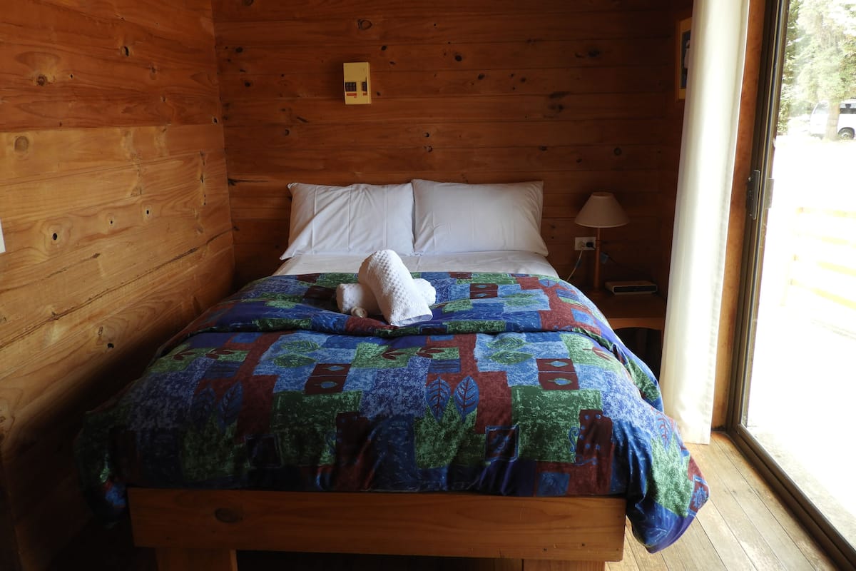 A wooden cabin room with a bed, white pillows, a quilt, a lamp, and a window.