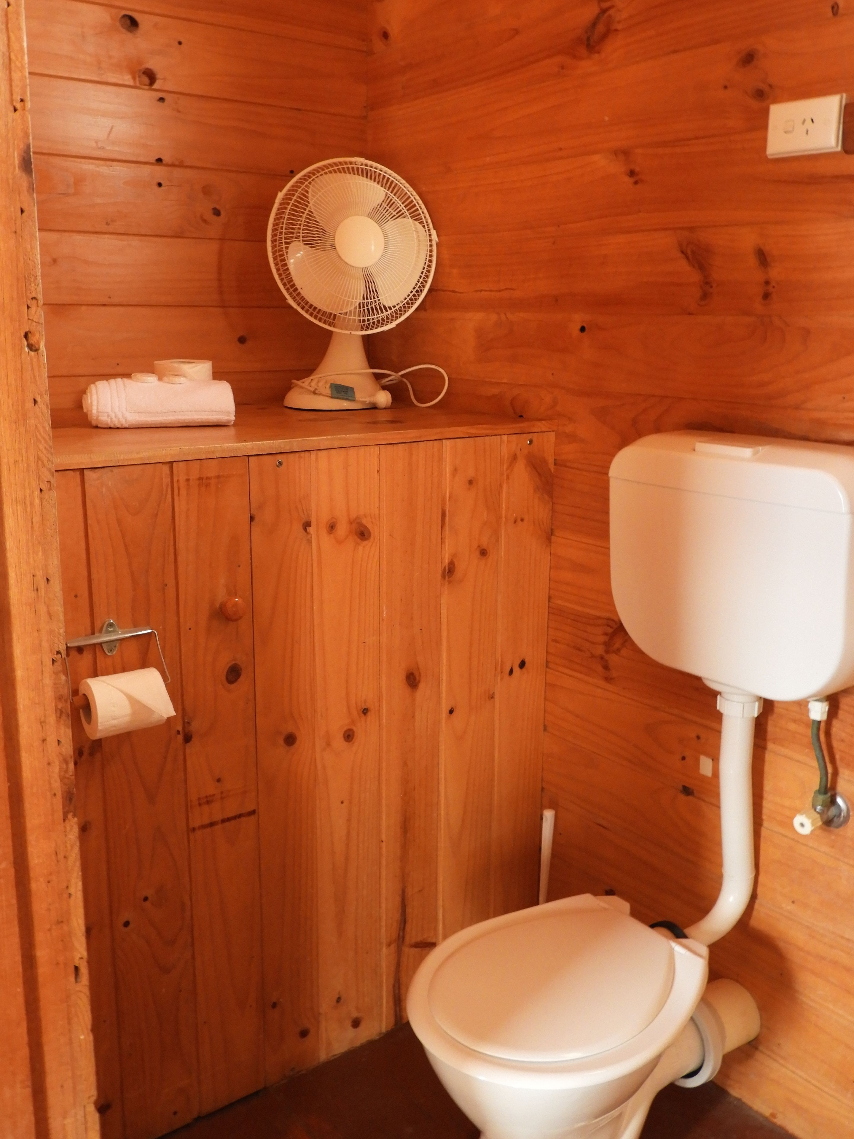 A wooden bathroom with a toilet, a fan, and a towel on a shelf.