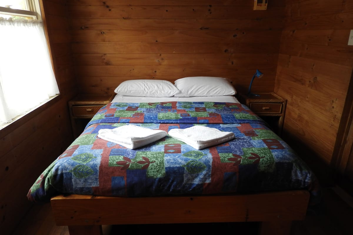 A wooden-paneled room with a bed, two white pillows, a blue and red quilt, and two towels on the bed.