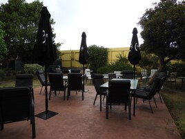 Um pátio externo com cadeiras e mesas pretas, guarda-sóis e uma parede amarela ao fundo.