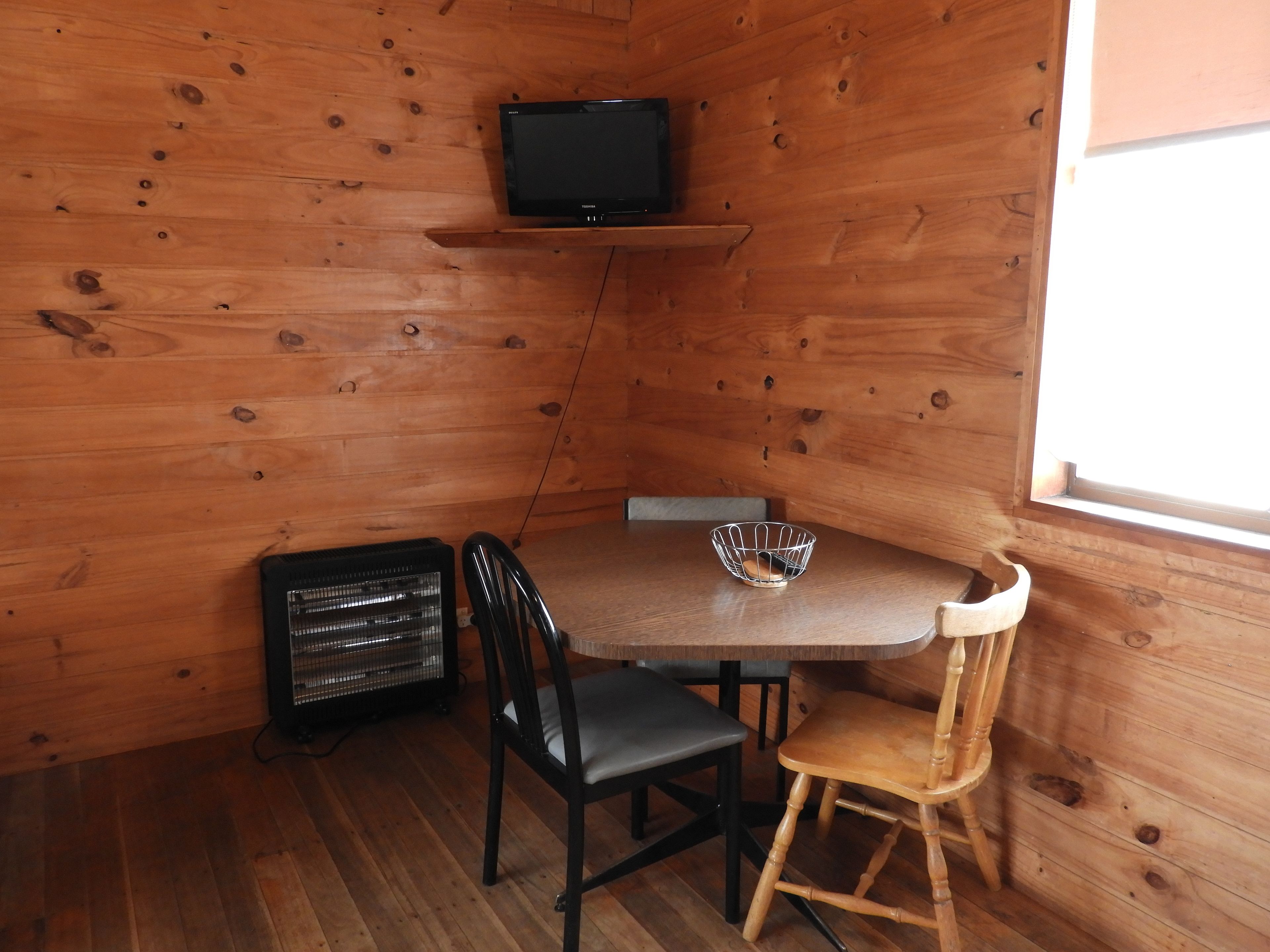 A wooden cabin interior with a TV on a shelf, a table with chairs, and a window.