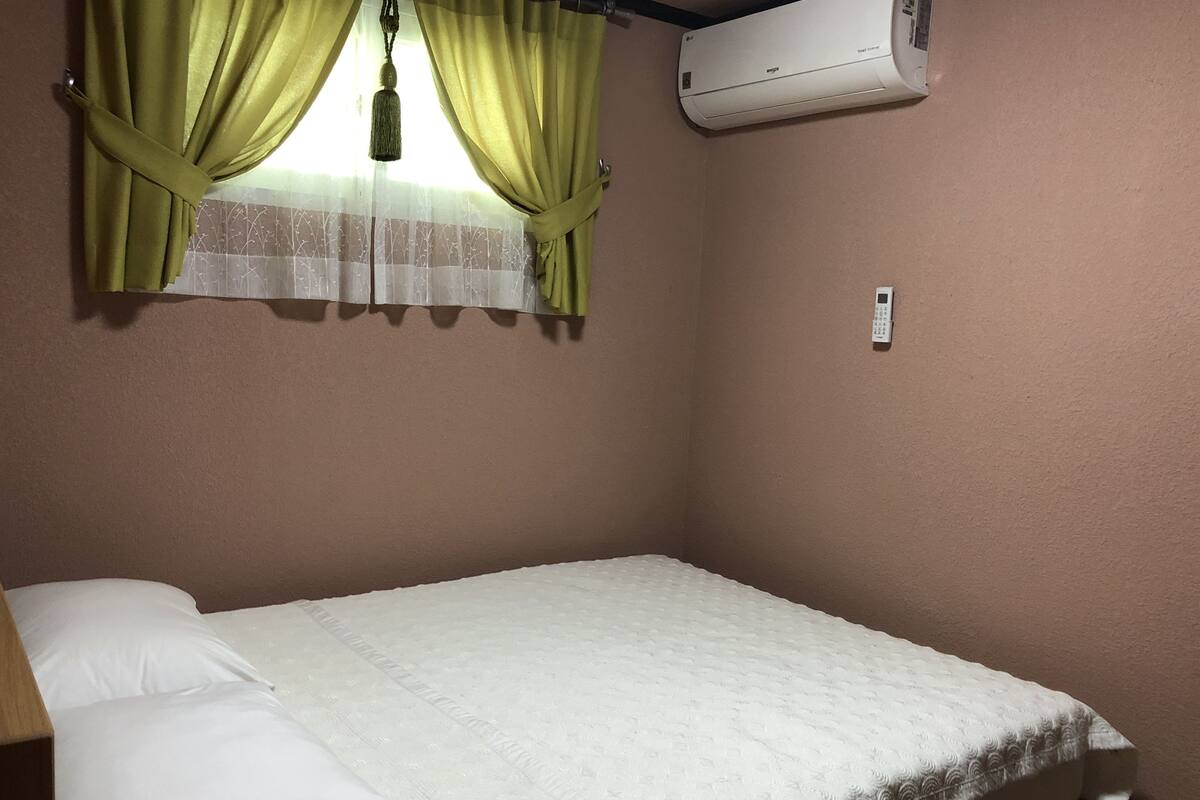 Un lit simple avec un matelas et des oreillers blancs, une fenêtre avec des rideaux verts et un appareil de climatisation fixé au mur.
