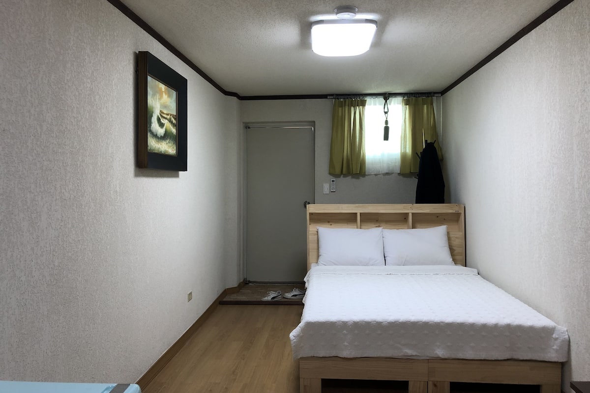 Une petite pièce simple, avec un lit, une porte, un luminaire et un tableau accroché au mur.