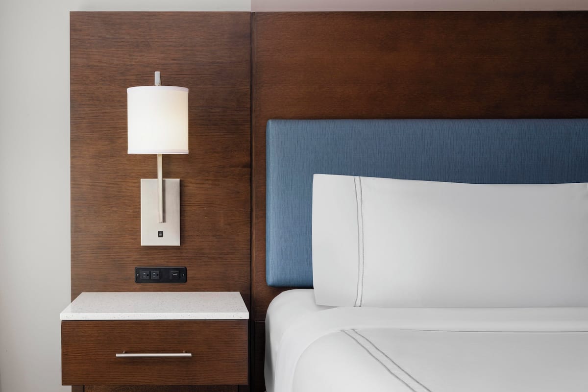A hotel room with a wooden headboard, a bedside table with a lamp, a drawer, and a bed with white bedding.