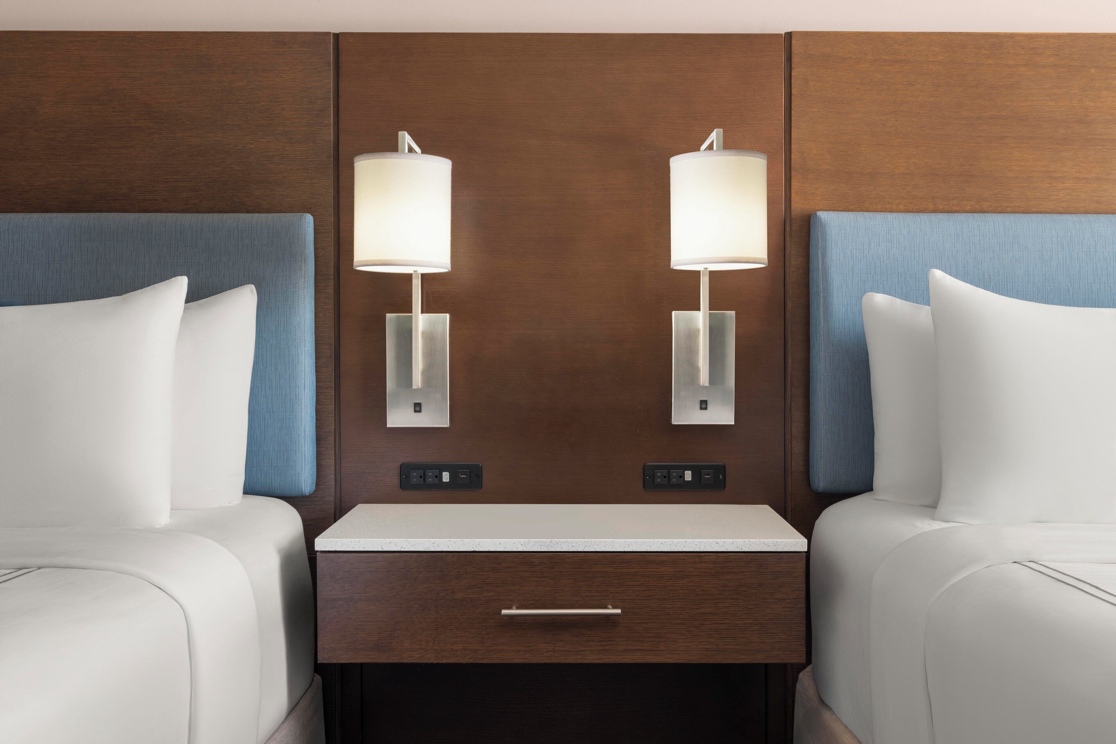 A hotel room with two beds, a nightstand with a drawer, and wall-mounted lamps.