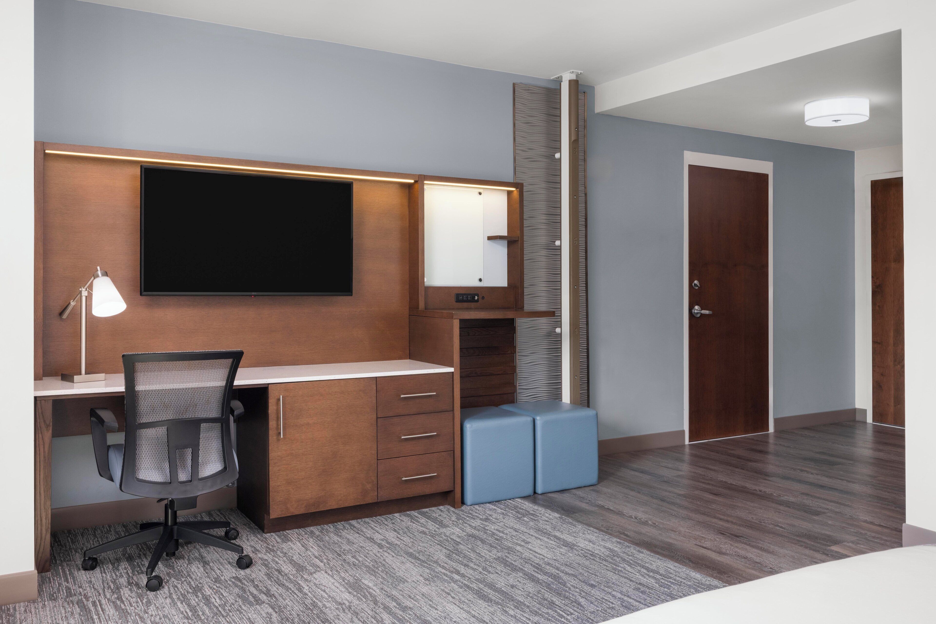 A modern hotel room with a wooden desk, a flat-screen TV, a desk lamp, a chair, and a large mirror.