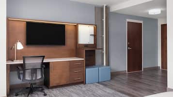 A modern hotel room with a wooden desk, a flat-screen TV, a desk lamp, a chair, and a large mirror.