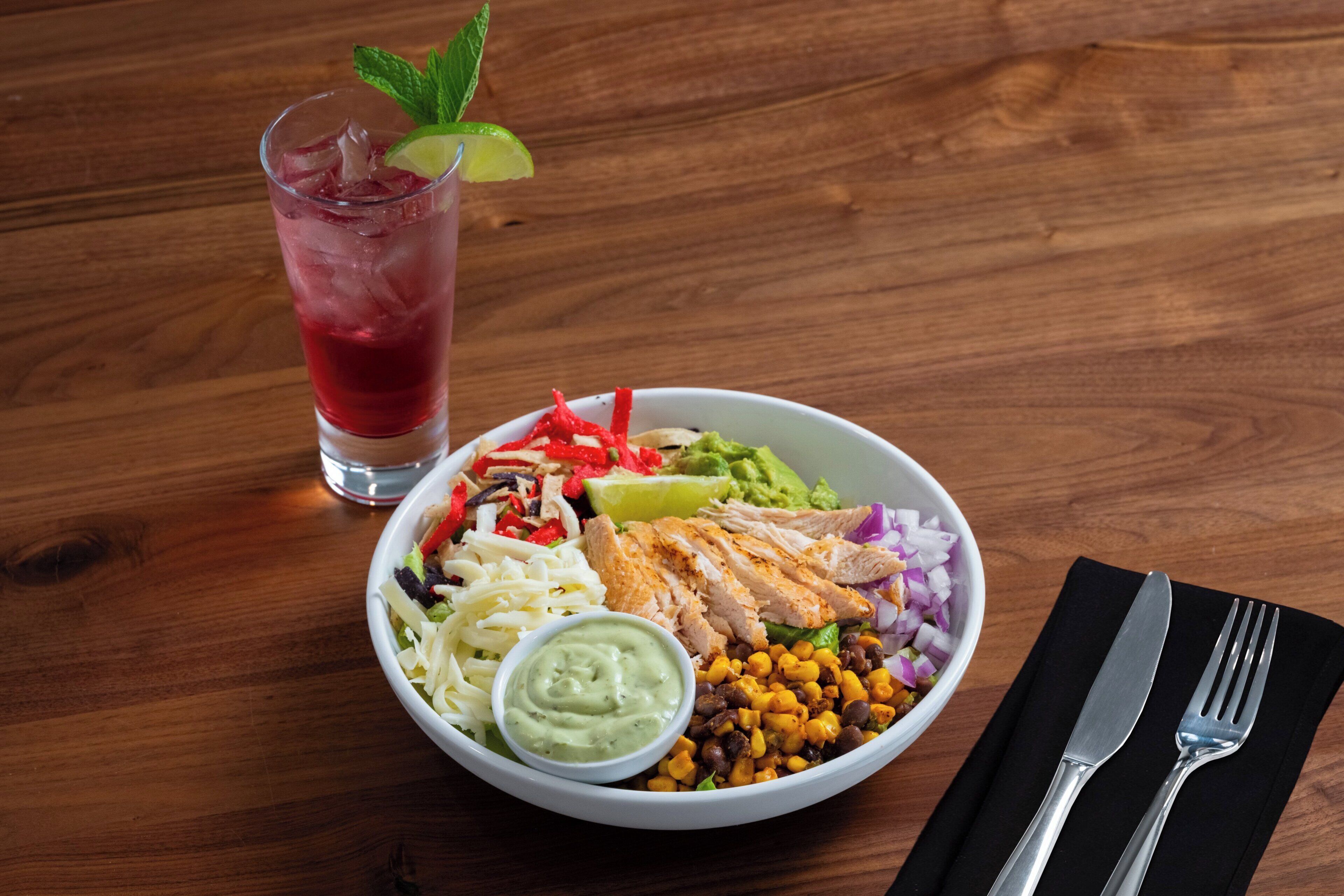 A meal consisting of grilled chicken, vegetables, and a green sauce, served with a glass of iced beverage on a wooden table.