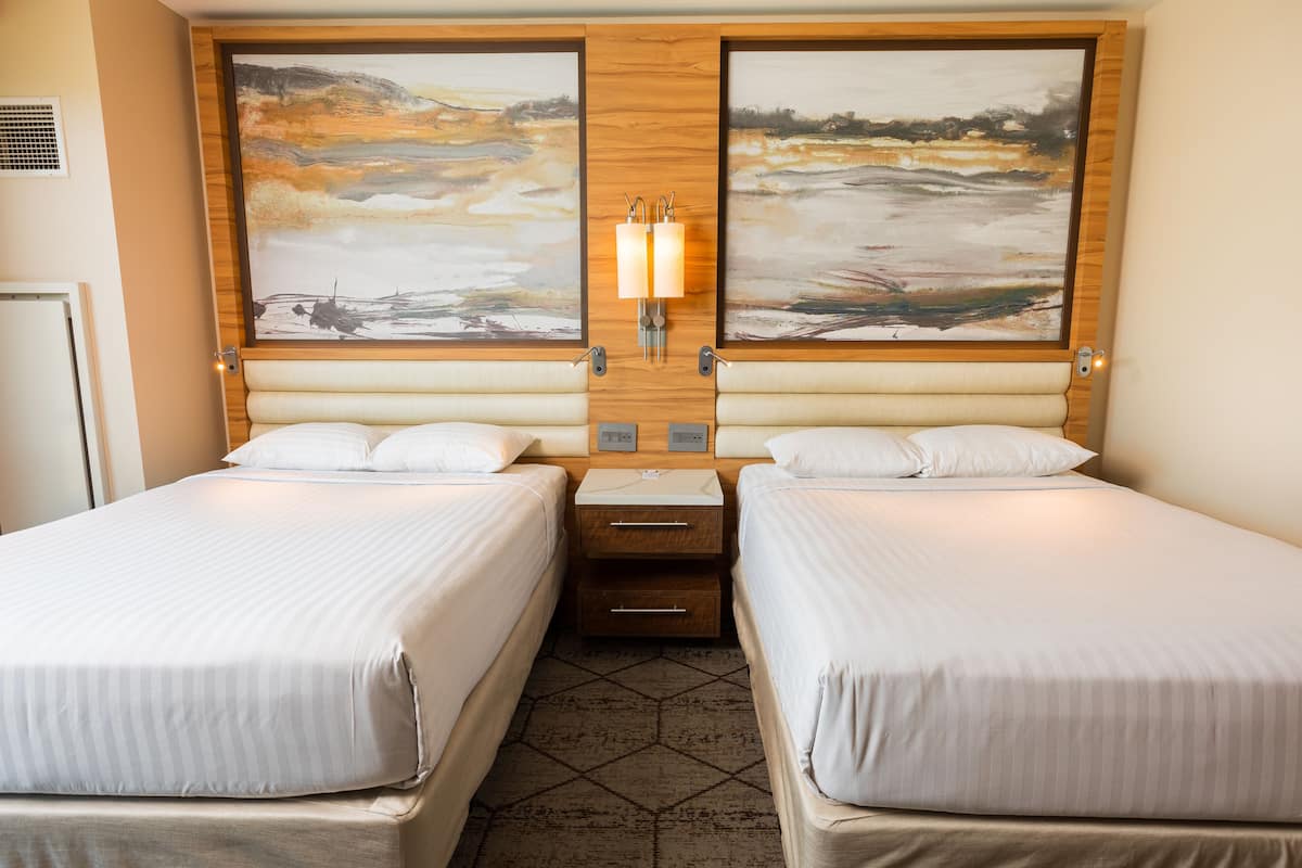 A hotel room with two beds, a wooden headboard, a nightstand, and a wall-mounted light fixture.