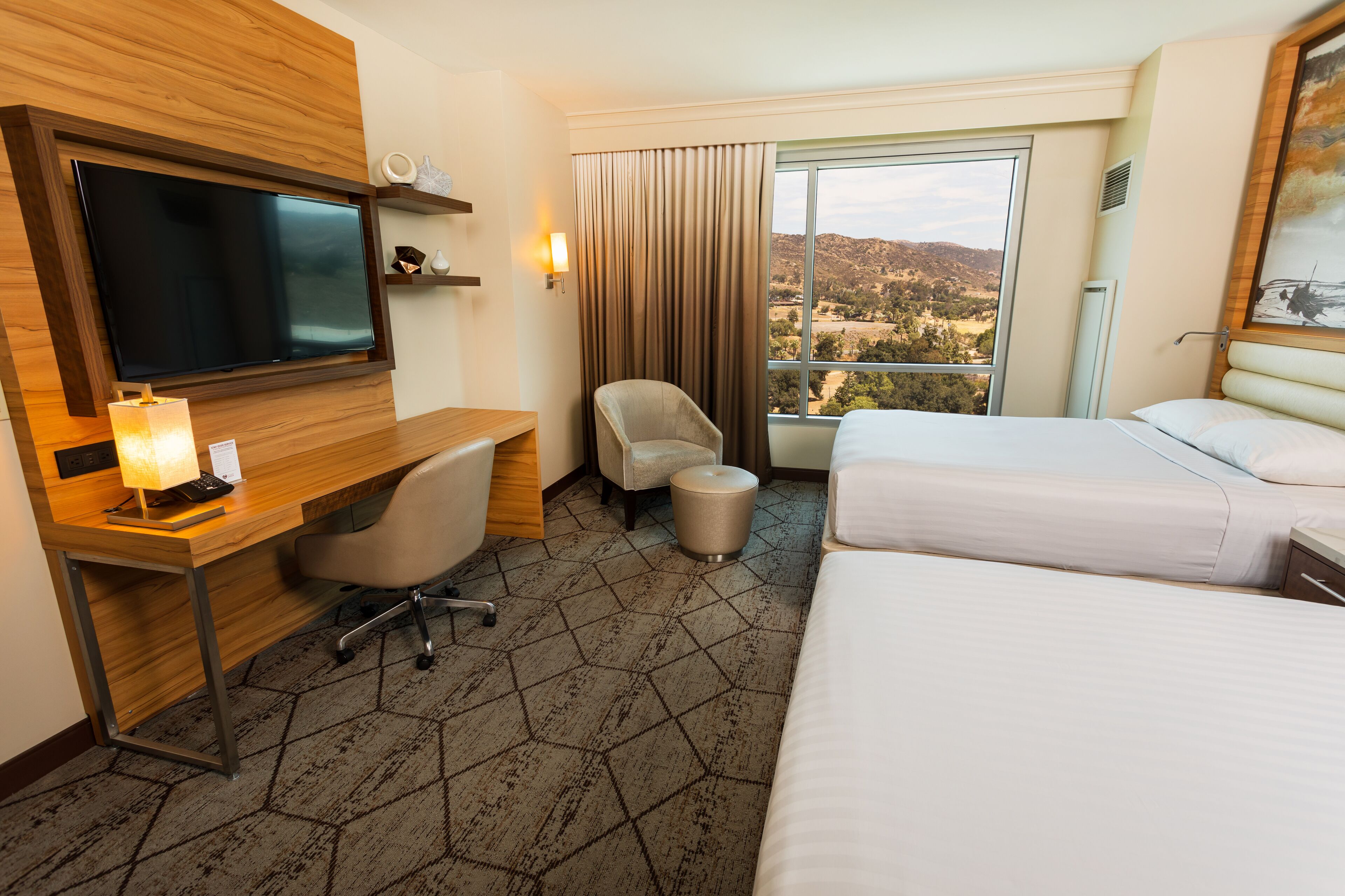 A hotel room with a desk, two beds, a chair, a TV, and a large window with a view.