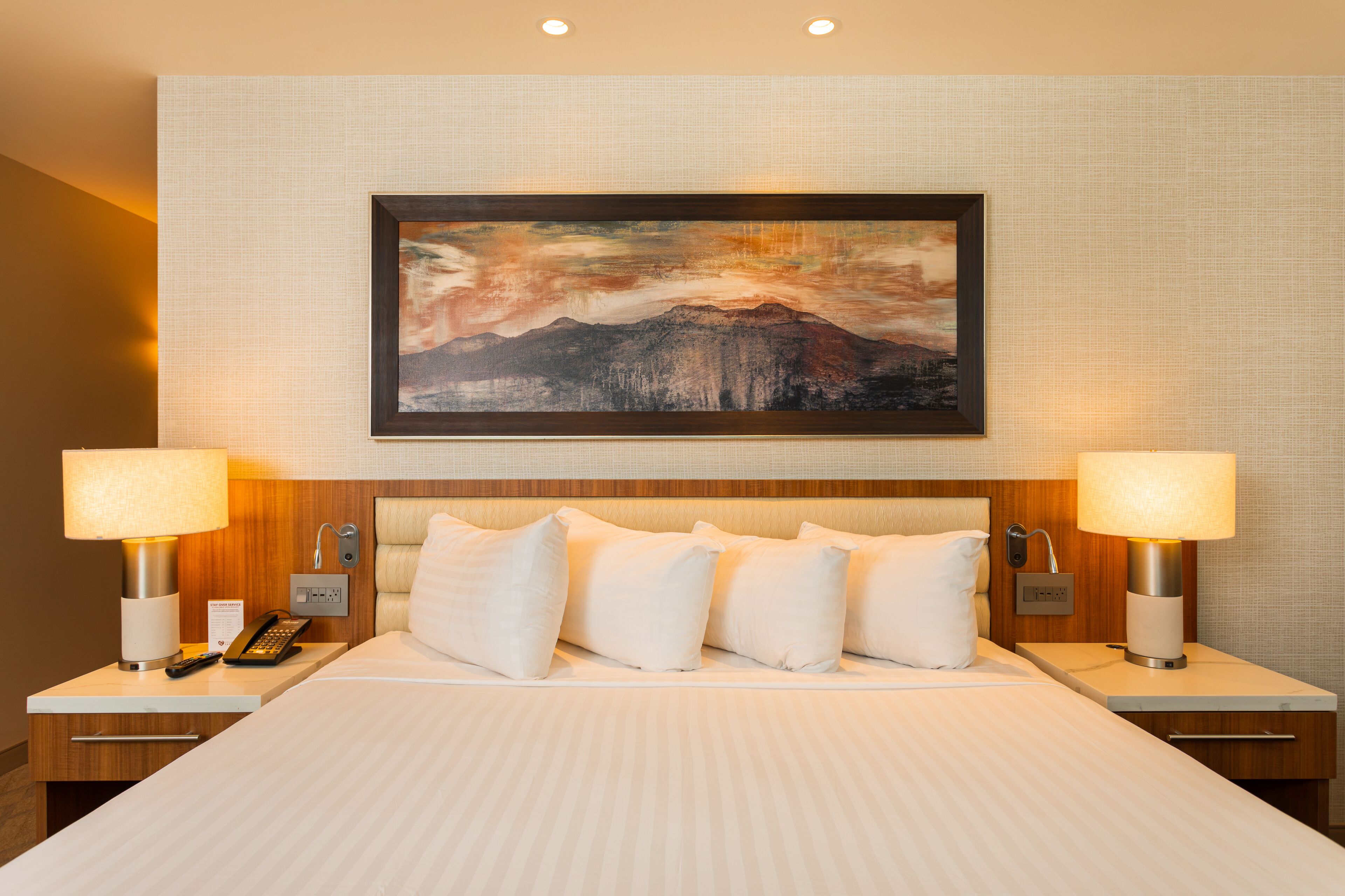 A hotel room with a large bed, two bedside tables, a mounted painting, and a nightstand with a lamp and phone.